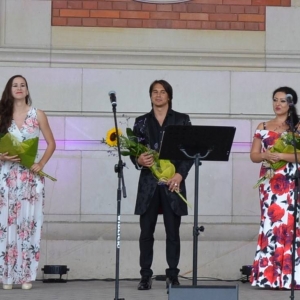 Zdjęcie przedstawia artystów na scenie amfiteatru w świerklanieckim parku z kwiatami po koncercie